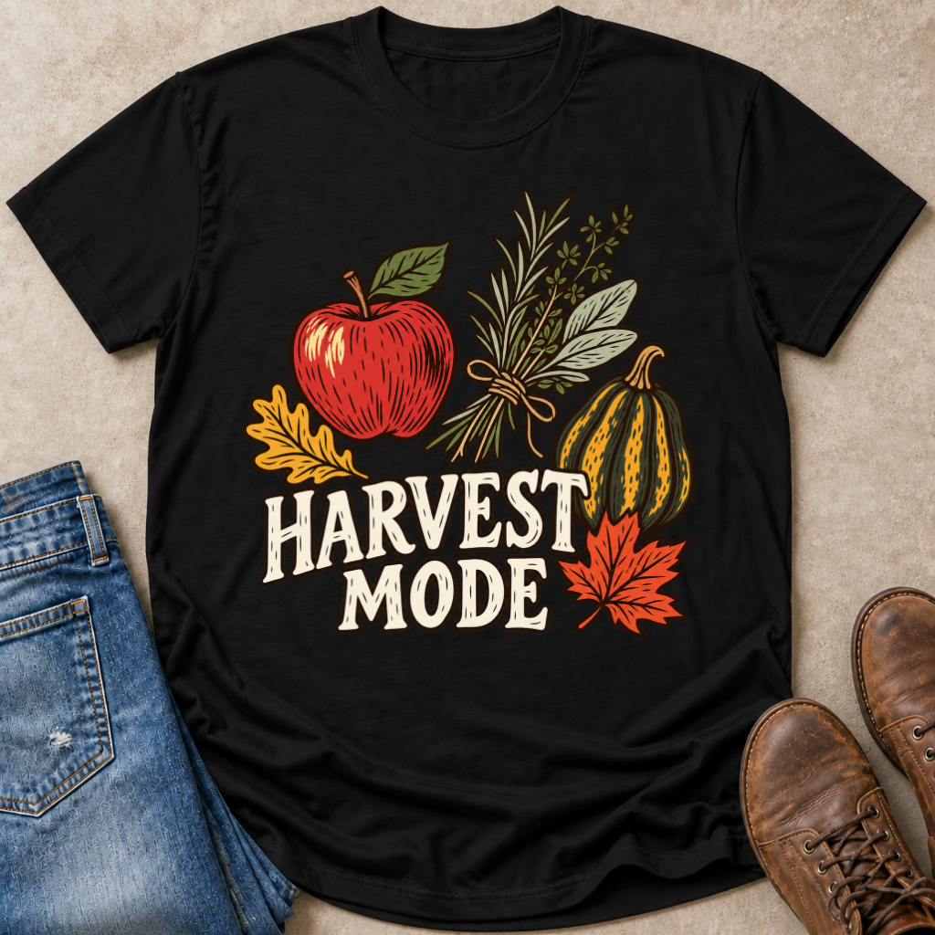 A black short-sleeve graphic T-shirt laid flat on a beige surface, styled with blue jeans, brown shoes, and autumn-themed illustrations including an apple, herbs, a gourd, leaves, and the words “HARVEST MODE.”.