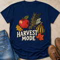 A navy blue graphic t-shirt laid flat on a beige surface, styled with blue jeans, brown boots, and autumn-themed illustrations including an apple, pumpkin, leaves, and the words 'HARVEST MODE'.