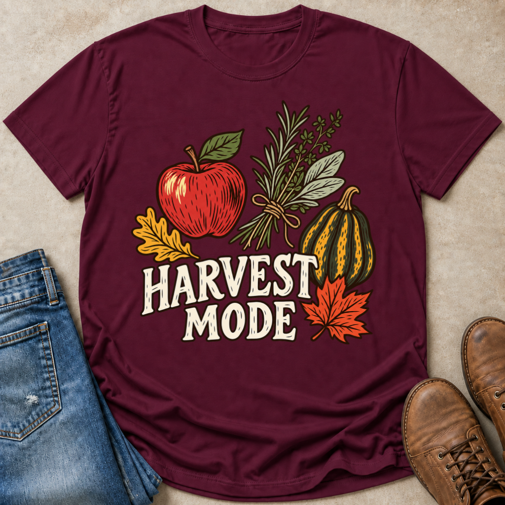 A maroon short-sleeve graphic T-shirt laid flat on a beige surface, styled with blue jeans, brown shoes, and autumn-themed illustrations including an apple, herbs, a gourd, leaves, and the words “HARVEST MODE.”.