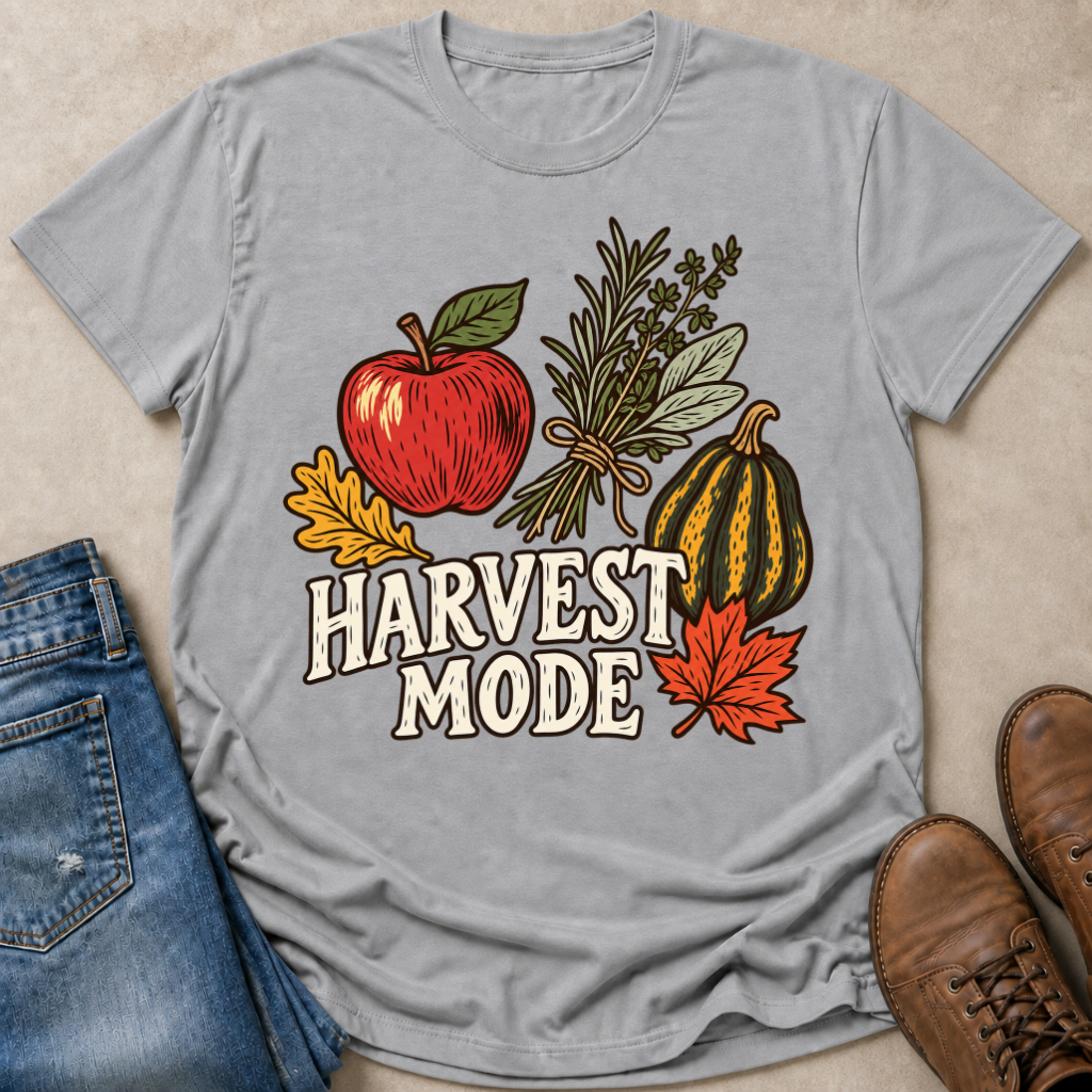 A light gray graphic t-shirt laid flat on a beige surface, styled with blue jeans, brown shoes, and autumn-themed illustrations including an apple, herbs, a gourd, and the words 'HARVEST MODE'.