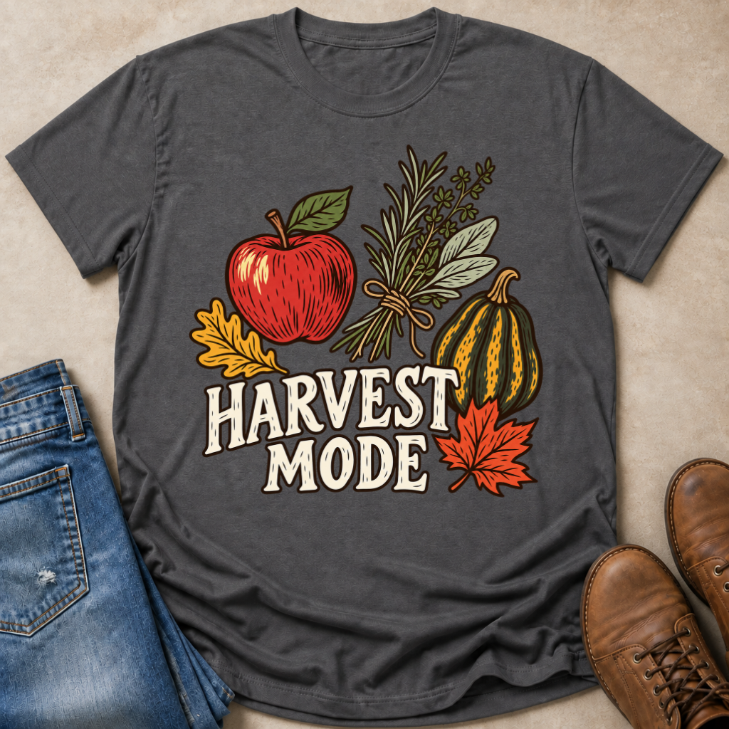 A dark gray graphic t-shirt laid flat on a beige surface, styled with blue jeans, brown boots, and autumn-themed illustrations including an apple, herbs, a pumpkin, leaves, and the words 'HARVEST MODE'.
