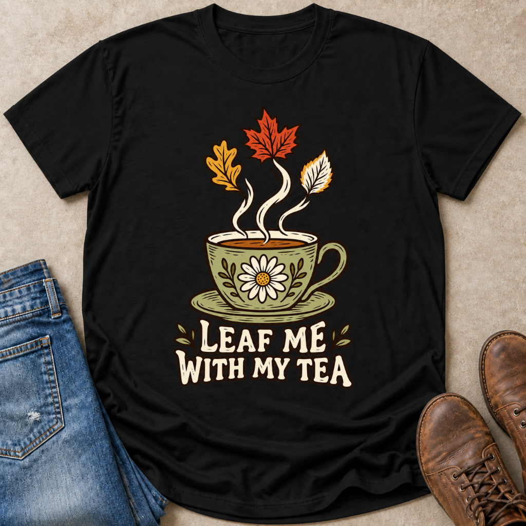 A black short-sleeve graphic T-shirt laid flat on a beige textured surface, styled with blue jeans, brown shoes, and a small autumn-themed tea cup illustration above the phrase “LEAF ME WITH MY TEA.”.