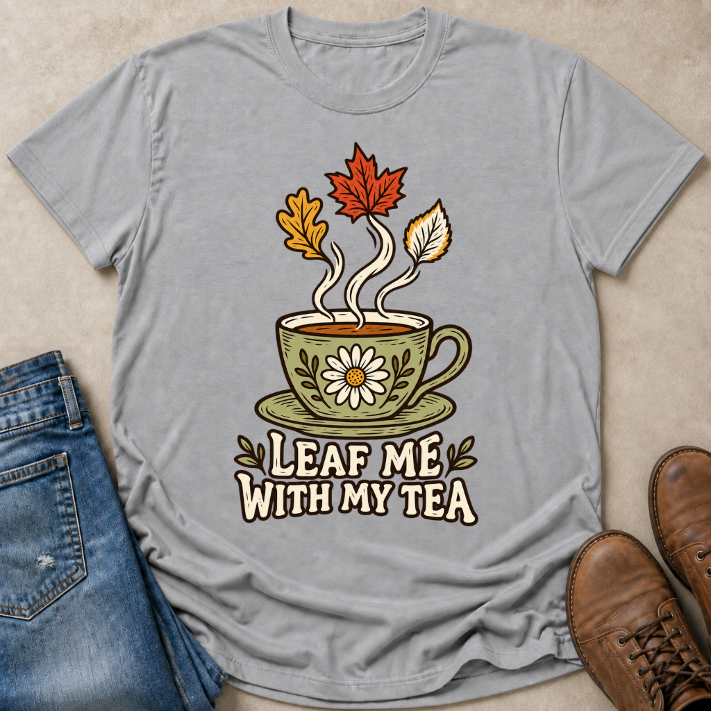 A light gray graphic t-shirt laid flat on a beige surface, styled with blue jeans and brown leather shoes. The t-shirt features a graphic of a green teacup with a daisy, steam, and autumn leaves rising from it, with the text 'LEAF ME WITH MY TEA' printed below.