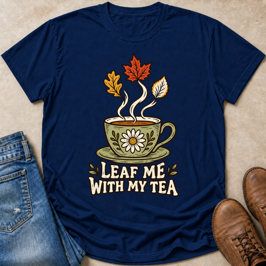 A navy blue graphic t-shirt laid flat on a beige surface, styled with blue jeans and brown leather boots. The t-shirt features a graphic of a green teacup with a daisy, steam, and autumn leaves rising from it, above the text 'LEAF ME WITH MY TEA'.