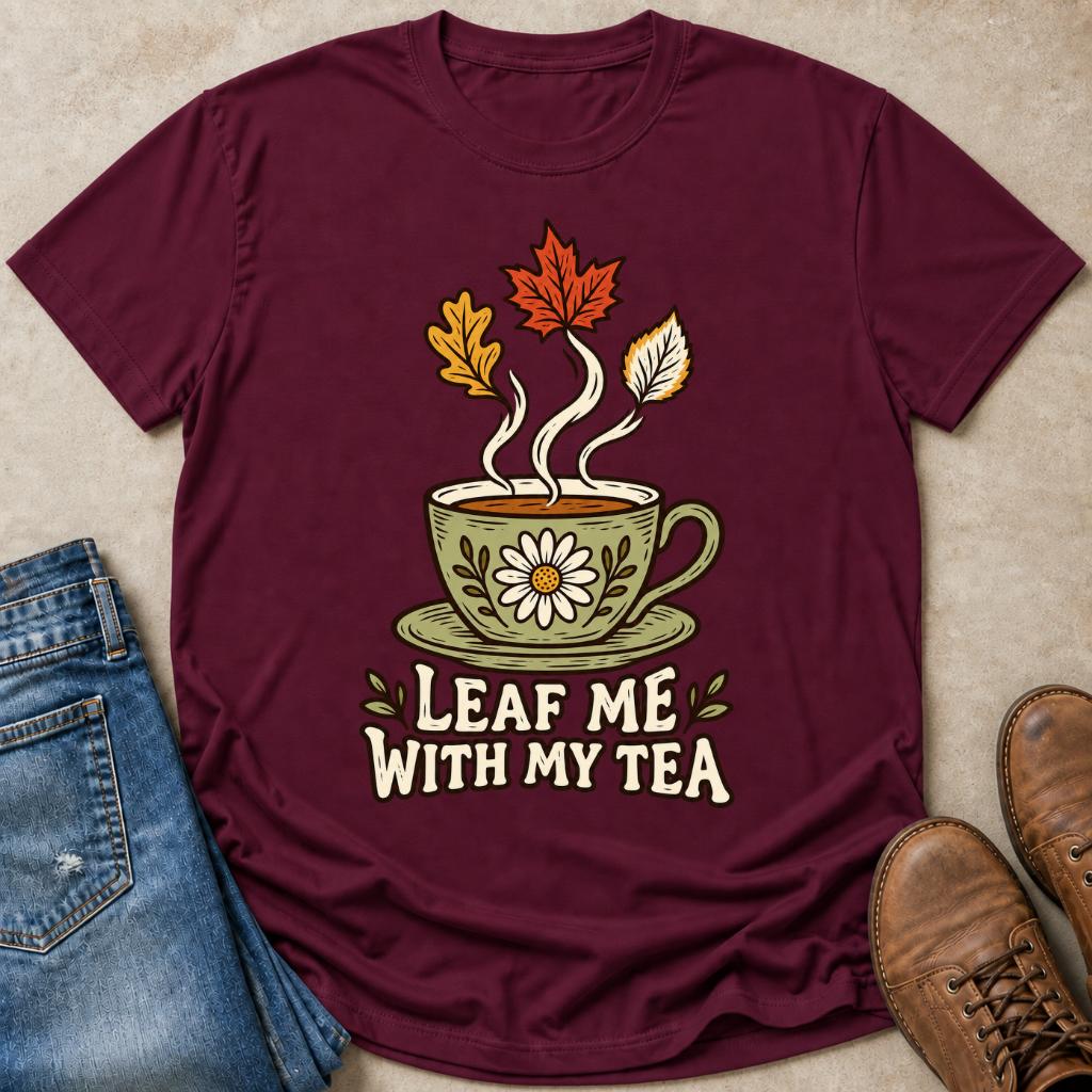 A maroon short-sleeve graphic T-shirt laid flat on a beige textured surface, styled with blue jeans on the left and brown boots on the right. The shirt features a central illustration of a green teacup with a daisy, steam, and autumn leaves above the phrase 'LEAF ME WITH MY TEA' in cream lettering.
