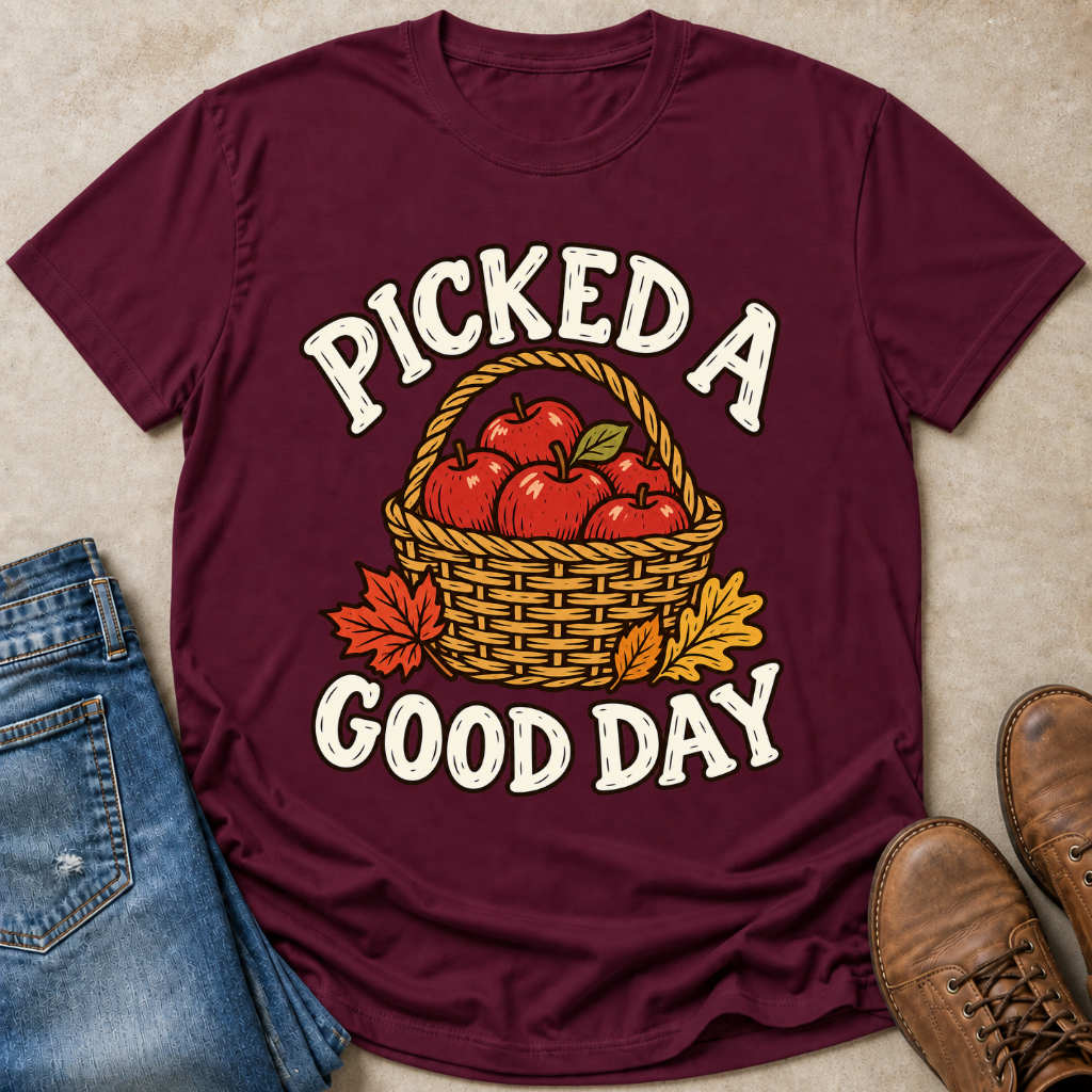 A maroon short-sleeve graphic T-shirt laid flat on a beige textured surface, styled with blue jeans, brown shoes, and autumn leaves. The shirt graphic reads “PICKED A GOOD DAY” around an illustration of red apples in a wicker basket.