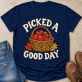 A navy blue graphic t-shirt laid flat on a beige surface, styled with blue jeans on the left and brown boots on the right. The t-shirt features a graphic of a wicker basket filled with red apples and autumn leaves, with the text 'PICKED A GOOD DAY' in white arched lettering.