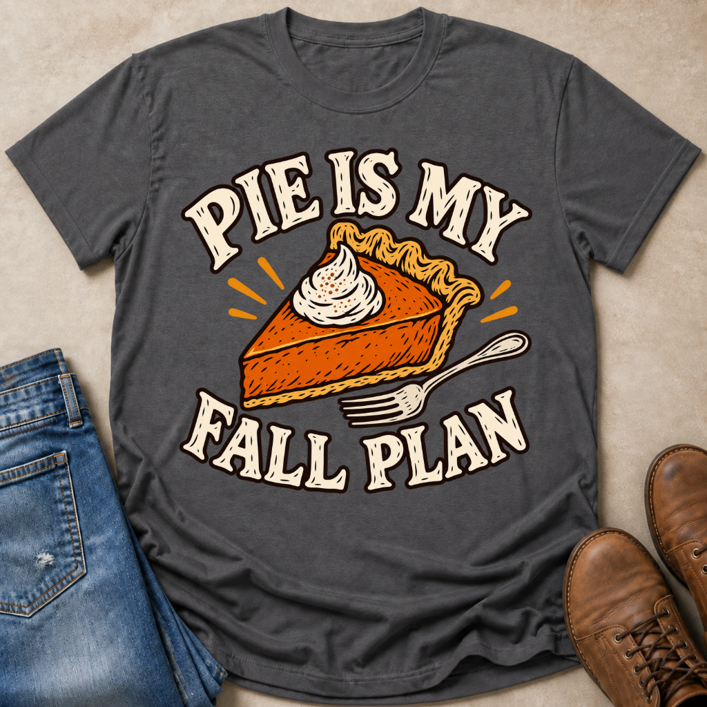 A dark gray graphic t-shirt laid flat on a beige surface, styled with blue jeans, brown boots, and orange accents. The t-shirt features a graphic of a slice of pumpkin pie with whipped cream and the text 'PIE IS MY FALL PLAN'.