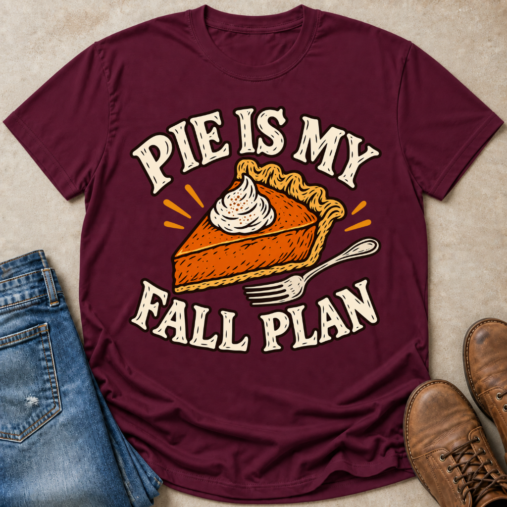 A maroon short-sleeve graphic T-shirt laid flat on a beige textured surface, styled with blue jeans, brown shoes, and small orange accents. The shirt graphic reads “PIE IS MY FALL PLAN” around an illustrated slice of pie with whipped topping and a fork.