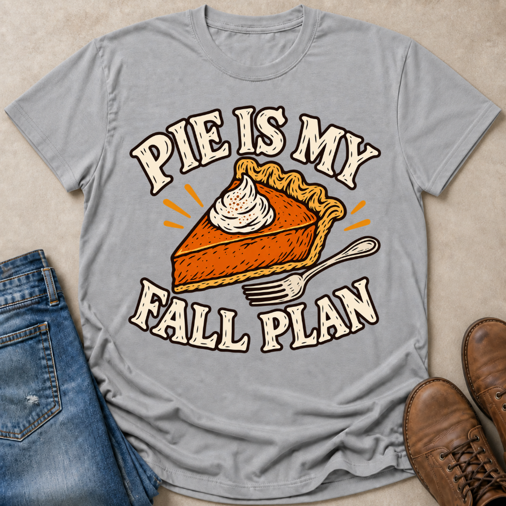 A light gray graphic t-shirt laid flat on a beige surface, styled with blue jeans on the left and brown leather shoes on the right. The t-shirt features a graphic of a slice of pumpkin pie with whipped cream and the text 'PIE IS MY FALL PLAN' in a retro cream-colored font.