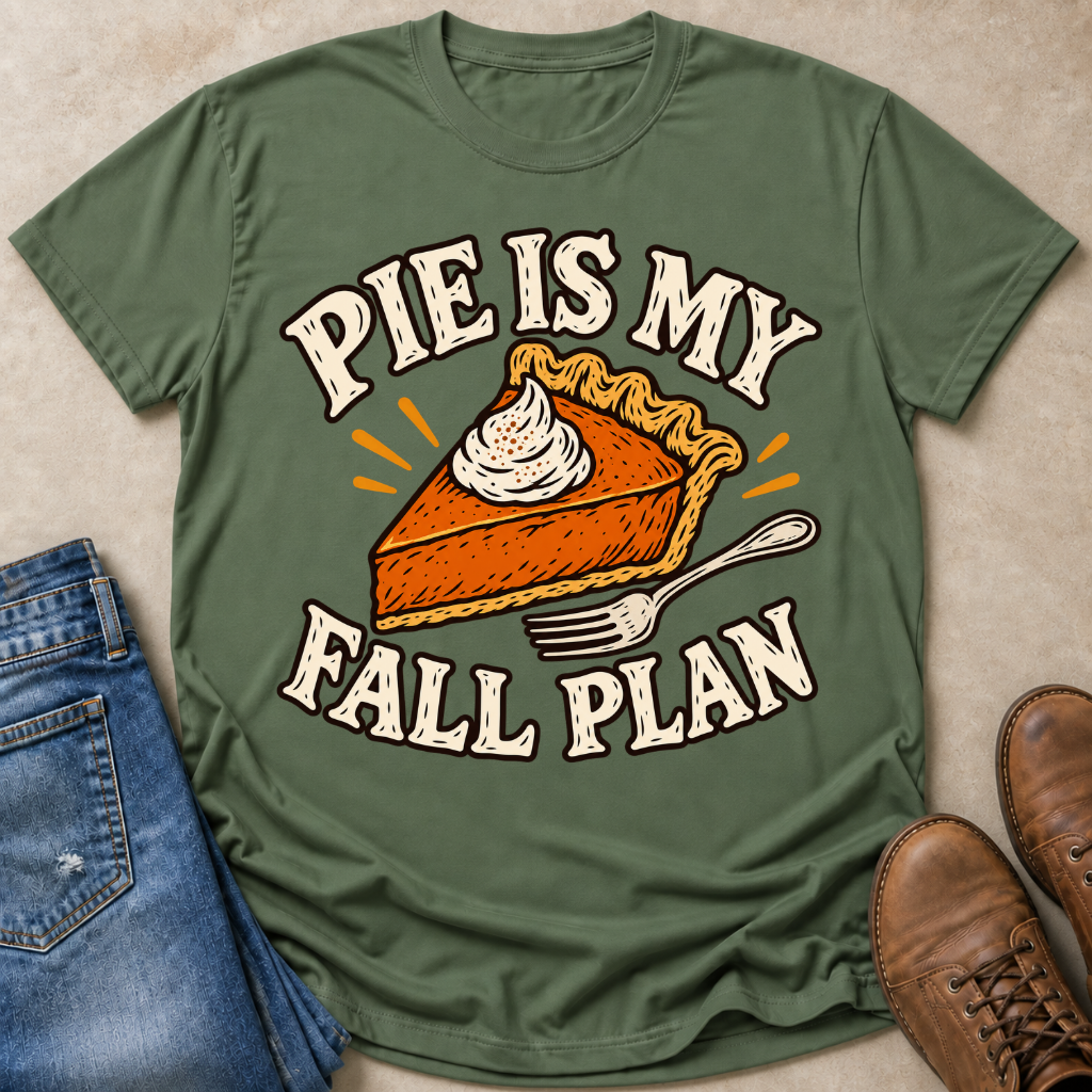 An olive green graphic t-shirt laid flat on a beige textured surface, styled with blue jeans, brown boots, and orange pumpkin pie slices. The t-shirt features a graphic of a slice of pumpkin pie with whipped cream and the text 'PIE IS MY FALL PLAN' in cream-colored letters with orange outlines.