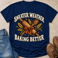 A navy blue graphic t-shirt laid flat on a beige surface, styled with blue jeans on the left and brown boots on the right. The t-shirt features a graphic with the text 'SWEATER WEATHER, BAKING BETTER' in white, surrounded by a rolling pin, wooden spoon, wheat stalks, and a red ribbon.