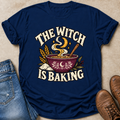 A navy blue graphic t-shirt laid flat on a beige textured surface, styled with blue jeans on the left and brown leather shoes on the right. The t-shirt features a graphic of a bowl of steaming baking mix with wheat stalks and the text 'THE WITCH IS BAKING' in cream and gold.