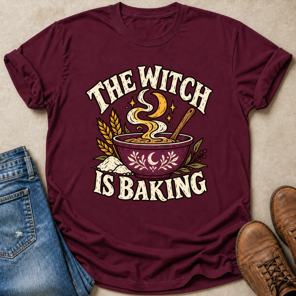 A maroon short-sleeve graphic T-shirt laid flat on a beige surface, styled with blue jeans on the left and brown boots on the right. The shirt features a large front print reading “THE WITCH IS BAKING” around an illustration of a bowl of stew with steam, wheat stalks, leaves, stars, and flour.