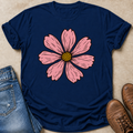 A navy blue short-sleeve t-shirt with a large pink cosmos flower graphic on the front, styled in a flat lay with blue jeans and brown boots on a beige textured background.