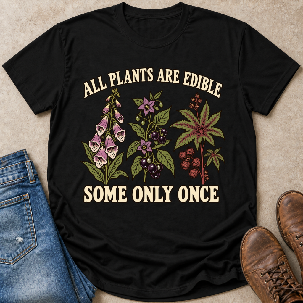 A black graphic t-shirt laid flat on a textured beige surface, styled with blue jeans on the left and brown leather shoes on the right. The t-shirt features a graphic of various toxic plants with the text 'ALL PLANTS ARE EDIBLE SOME ONLY ONCE' in a cream-colored font.